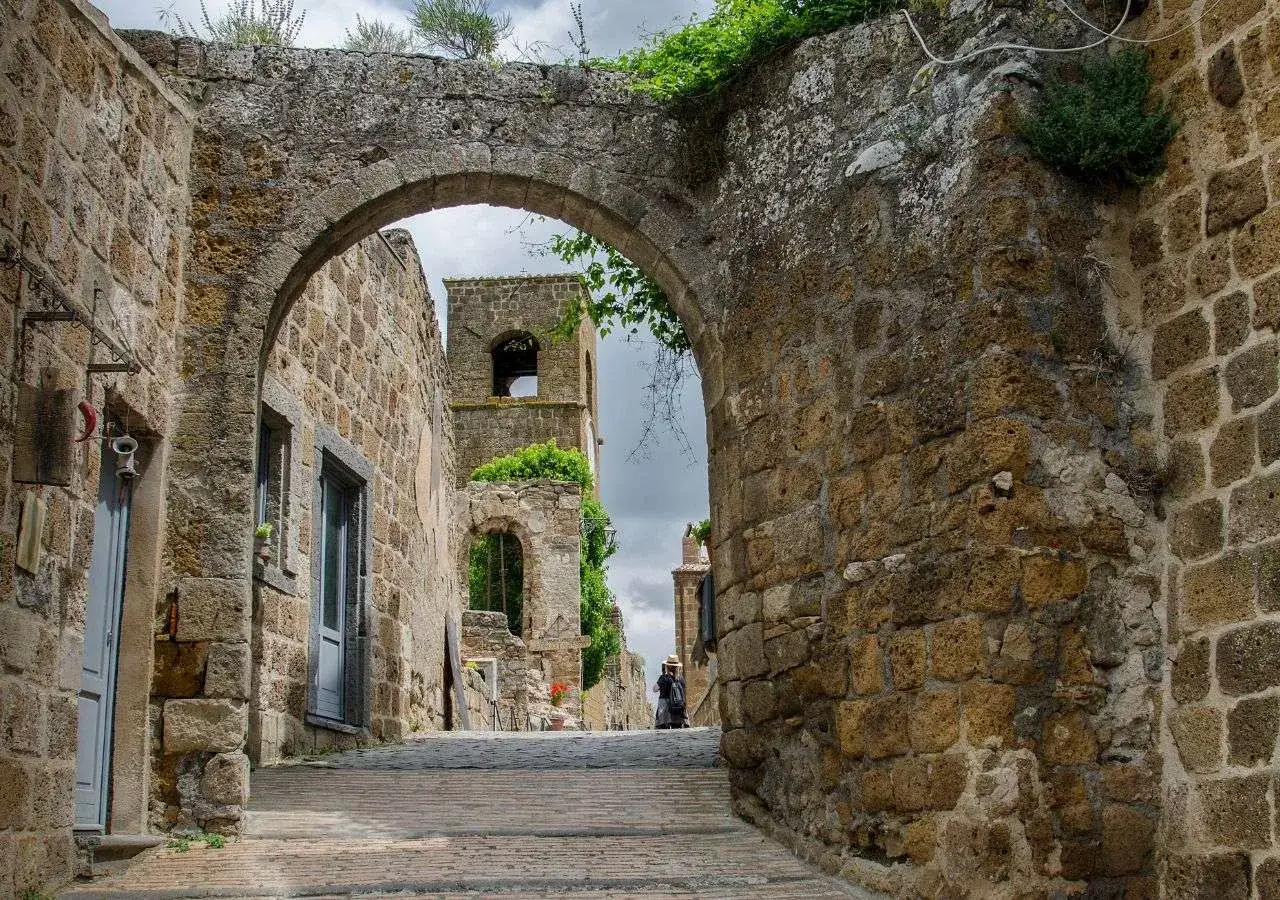 Un'antica via lastricata conduce a un arco di pietra, oltre il quale si intravede una torre in rovina. Un'atmosfera da città fantasma nel Lazio, con vegetazione che cresce tra le pietre.