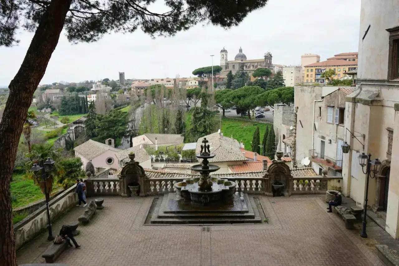 Vista panoramica di un borgo con fontana centrale, edifici storici e il Palazzo Priori in lontananza.