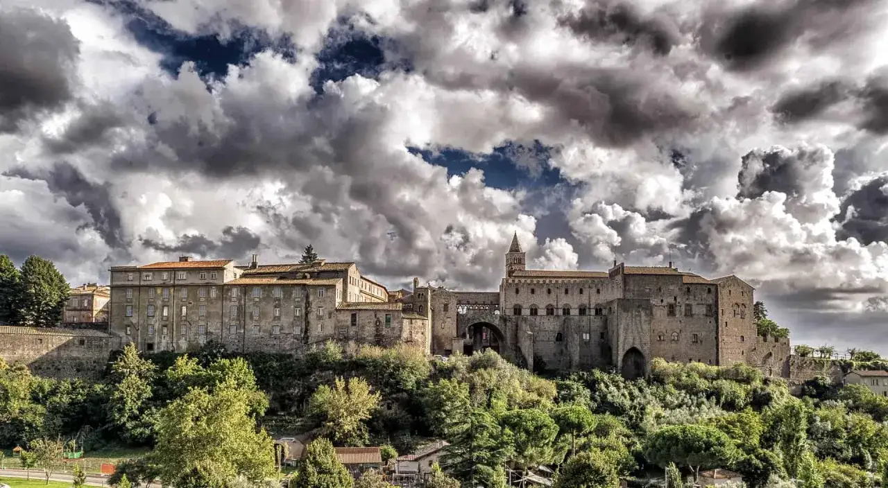 Antico borgo medievale in Tuscia, dove si trova questo imponente complesso di edifici storici su una collina verdeggiante, sotto un cielo drammatico.