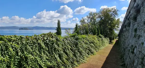 Un sentiero fiancheggiato da un muro di pietra e una siepe rigogliosa conduce alla vista del **lago di Capodimonte**, con un'isola in lontananza sotto un cielo azzurro e nuvoloso.