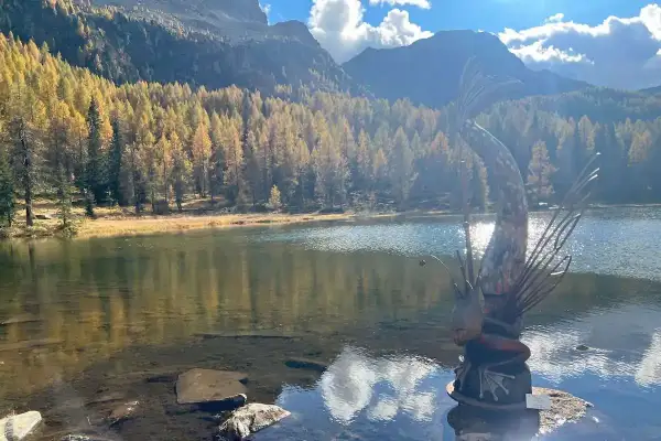 Scultura di un pesce con canna da pesca sul **lago di San Pellegrino**, circondato da boschi autunnali e montagne.