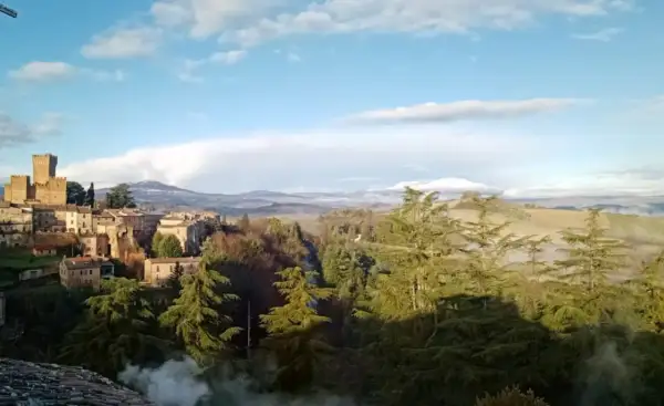Borgo medievale con torre imponente, immerso nel verde del comune di Proceno, con colline nebbiose all'orizzonte.