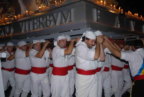 Uomini con fasce rosse e copricapi bianchi trasportano un pesante manufatto illuminato da candele. La scritta "VITERBVM" è visibile. La macchina di Santa Rosa caduta è un evento drammatico.