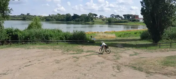 Ciclista pedala vicino al lago di Monterosi, con vegetazione rigogliosa e cielo nuvoloso.